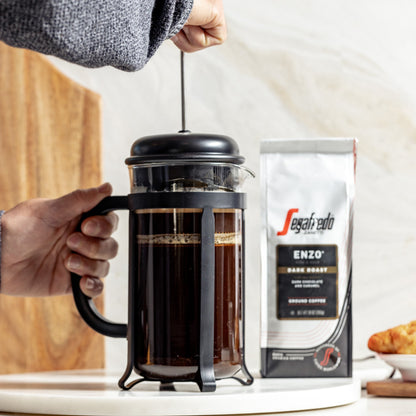 A hand presses a French press filled with coffee beside an Enzo Dark Roast bag from Segafredo Zanetti® Coffee, celebrating Italian tradition on the counter.